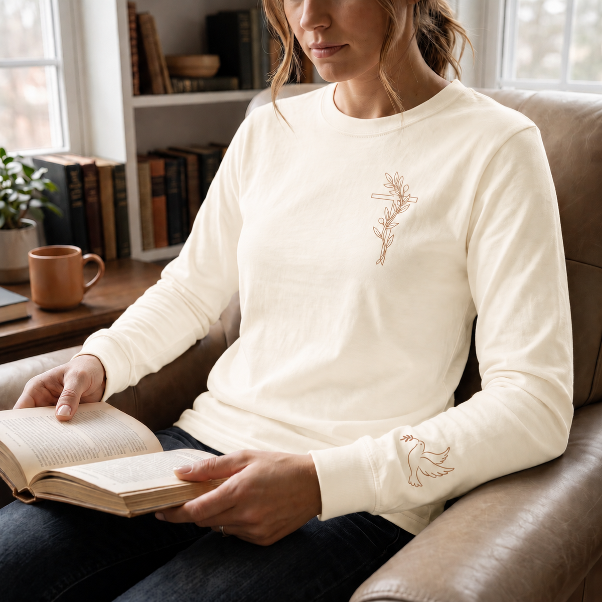Model wearing the ivory Olive Branch Cross & Dove long-sleeve tee while reading in a leather armchair; front cross and sleeve dove visible