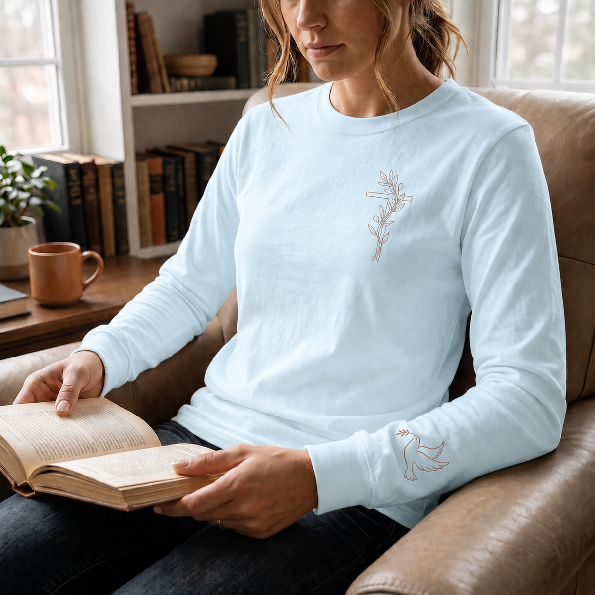 Model wearing the chambray Olive Branch Cross & Dove long-sleeve tee while reading in a leather armchair; front cross and sleeve dove visible