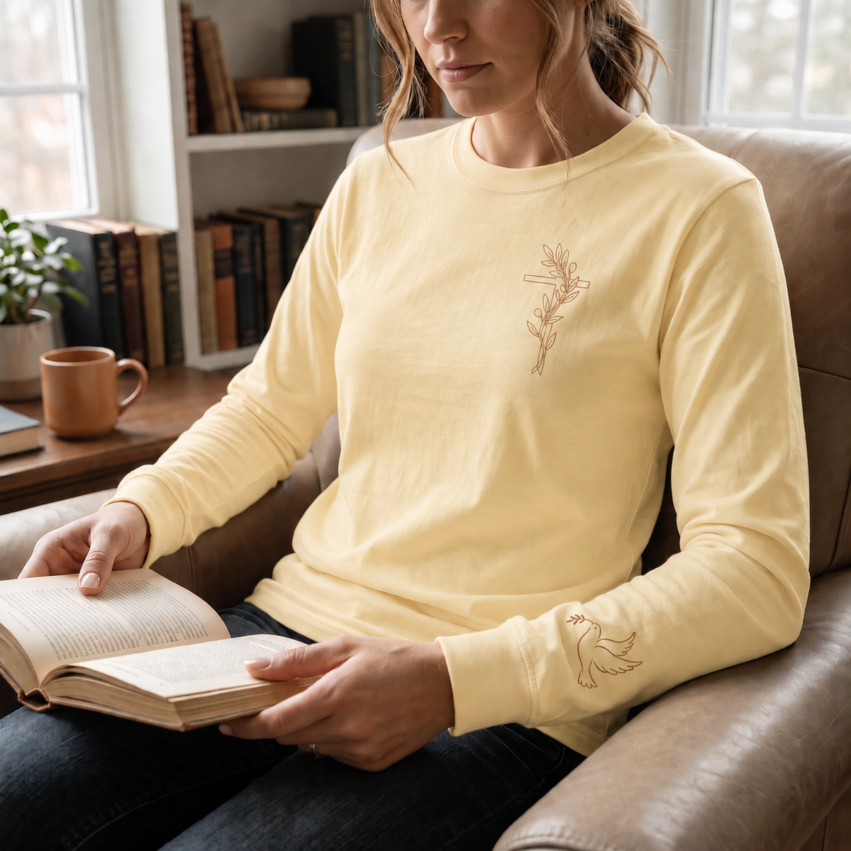Model wearing the butter Olive Branch Cross & Dove long-sleeve tee while reading in a leather armchair; front cross and sleeve dove visible
