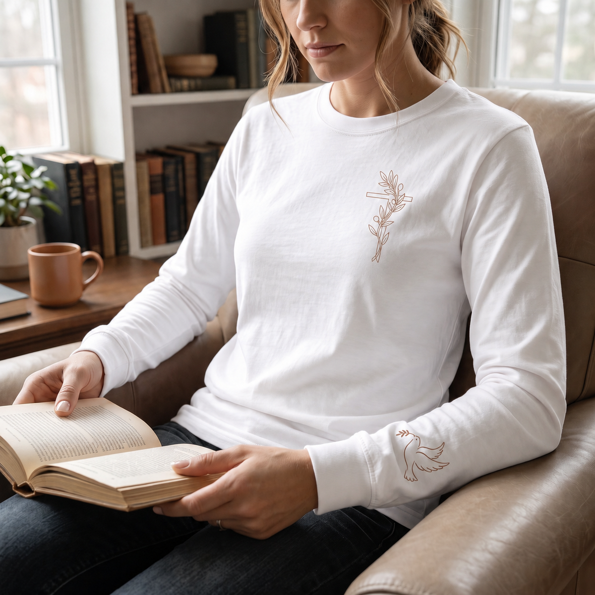 Model wearing the white Olive Branch Cross & Dove long-sleeve tee while reading in a leather armchair; front cross and sleeve dove visible
