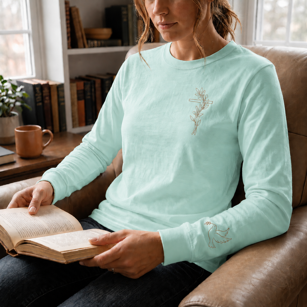 Model wearing the chalky mint Olive Branch Cross & Dove long-sleeve tee while reading in a leather armchair; front cross and sleeve dove visible