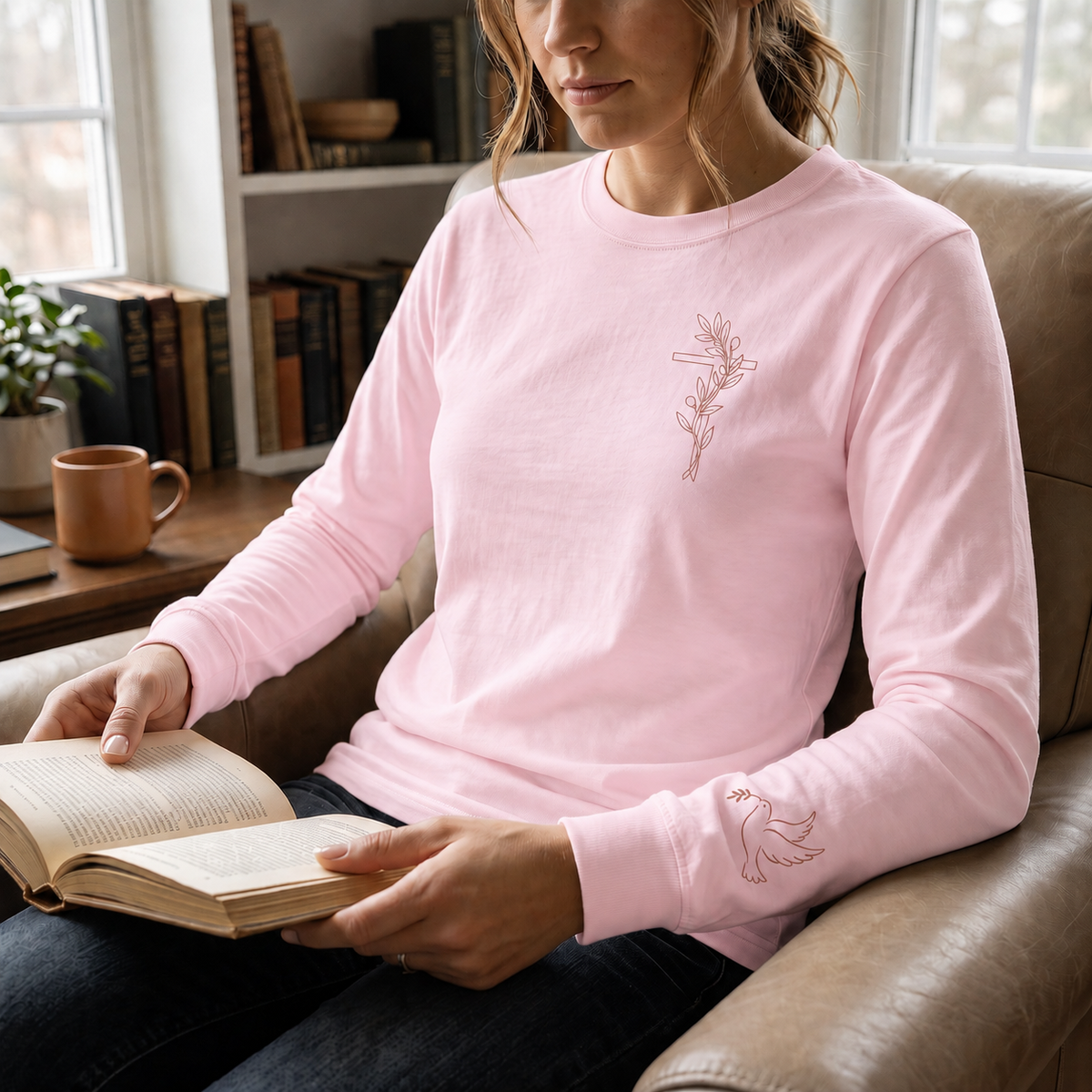 Model wearing the blossom Olive Branch Cross & Dove long-sleeve tee while reading in a leather armchair; front cross and sleeve dove visible