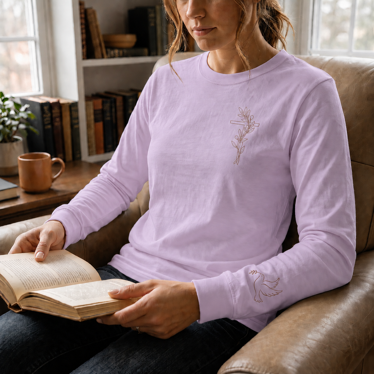 Model wearing the orchid Olive Branch Cross & Dove long-sleeve tee while reading in a leather armchair; front cross and sleeve dove visible
