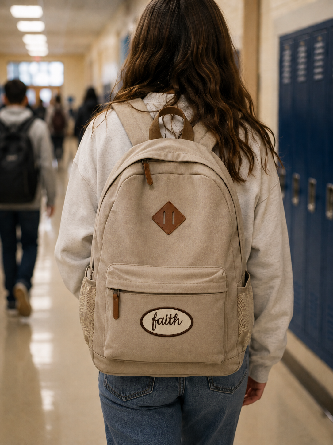 A person viewed from behind walking down a school hallway wearing a beige canvas backpack with brown leather-like trim and a front pocket featuring an oval patch that reads 'faith'.