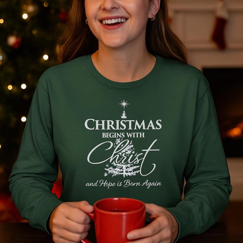 Person wearing a green sweatshirt with a Christmas message, holding a red mug in front of a decorated Christmas tree.