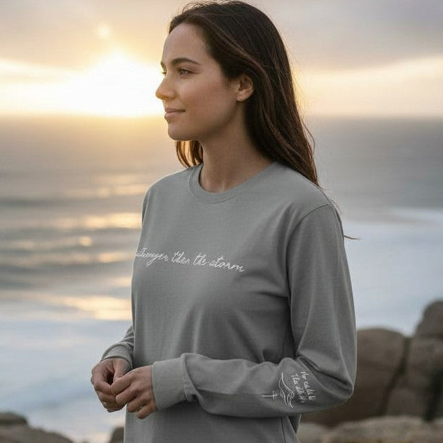 Woman wearing a gray long-sleeve shirt with text, standing by the ocean at sunset.
