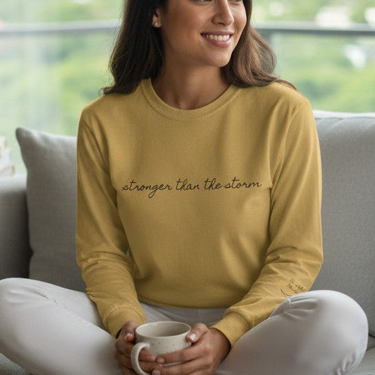 Woman sitting on a couch holding a mug, wearing a mustard yellow sweatshirt with text, in a room with large windows.
