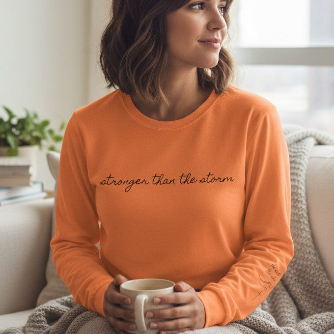 Woman in an orange sweater holding a mug, sitting on a couch in a bright room.