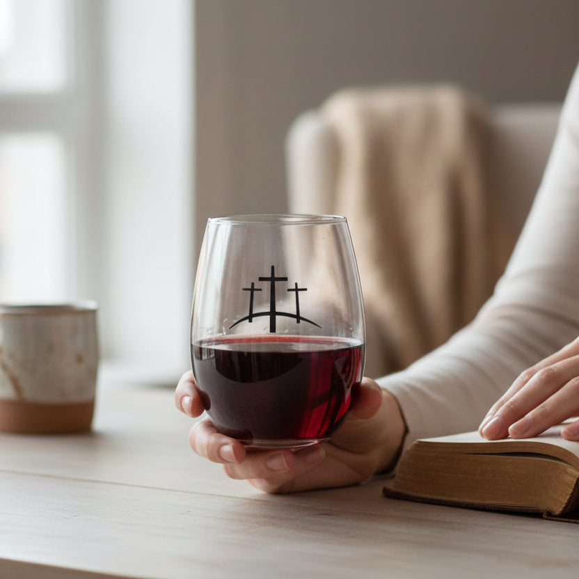Person holding a glass of red wine with a cross design, sitting at a table with a book and mug.